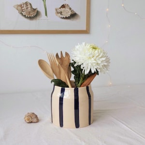 May include: A cylindrical ceramic utensil holder with a cream-colored base and vertical navy blue stripes. Inside the holder are several wooden kitchen utensils, including a spoon and fork, alongside a large, white, multi-petaled chrysanthemum flower.