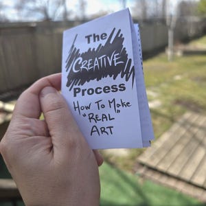May include: A small, folded booklet titled "The Creative Process" is held in a hand. The cover features the title in a mix of stylized and handwritten fonts, with the subtitle "How To Make 'REAL' ART". The background is a blurred outdoor scene.