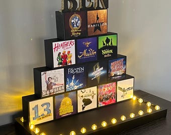 Puede incluir: Un expositor negro escalonado con el nombre BEN en letras doradas en la parte superior. El soporte contiene bloques cuadrados impresos con varios logotipos de musicales como Hamilton, Aladdin, Shrek, Frozen, Newsies y otros, iluminados por luces.