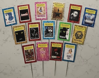 May include: A set of 12 colorful Broadway show themed cake toppers. Each topper features a different Broadway show logo and title, including Hairspray, Annie, Waitress, Hamilton, Phantom of the Opera, Dear Evan Hansen, Cats, Beauty and the Beast, Les Miserables, Wicked, The Lion King, Mean Girls, Hadestown, and Mamma Mia.