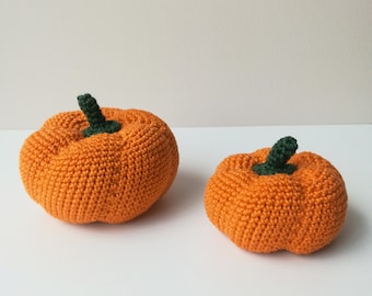 May include: Two crocheted pumpkins, one larger than the other, are displayed on a white surface. The pumpkins are orange with green stems.