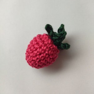 Peut inclure: Une fraise en crochet avec une tige et des feuilles vertes. La fraise est de couleur rose vif et les feuilles sont d'un vert foncé.