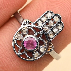 Peut inclure: Une bague en argent avec un motif de main de Hamsa. La bague est sertie de petits diamants blancs et d'une pierre précieuse rose au centre.