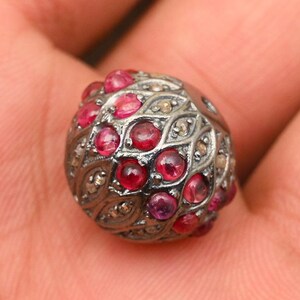 Könnte beinhalten: Eine runde silberne Perle mit einem schwarzen Metalldesign und roten und klaren Edelsteinen.