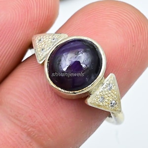 Może przedstawiać: Srebrny pierścionek z dużym, okrągłym, ciemnofioletowym kamieniem szlachetnym. Pierścionek ma dwa małe, trójkątne akcenty po obu stronach kamienia szlachetnego, każdy inkrustowany małymi, przezroczystymi kamieniami. Widoczny jest napis "shivanijewels".