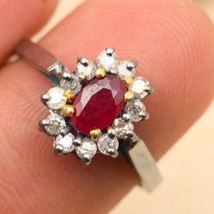 May include: A silver ring featuring a red oval gemstone encircled by a halo of smaller white gemstones.