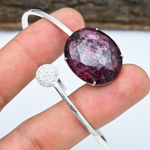 Puede incluir: Un brazalete plateado con una gran gema ovalada de color púrpura intenso. El diseño abierto incluye un acento circular más pequeño con diamantes. La gema es el punto focal, con una superficie facetada.