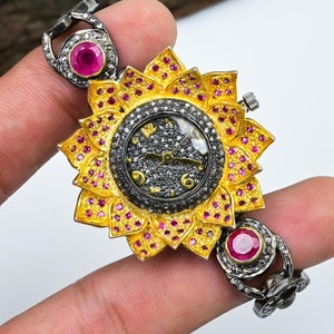 Puede incluir: Un reloj dorado y negro con un diseño de girasol. La esfera del reloj está rodeada de pequeñas piedras preciosas rosas y la correa está hecha de piezas de metal enlazadas con piedras preciosas rosas.