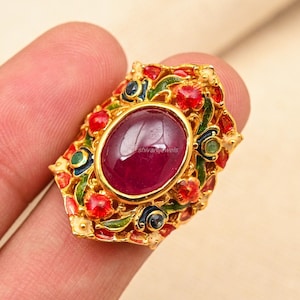 Puede incluir: Un anillo de oro con una gran piedra preciosa roja rodeada de pequeñas piedras preciosas verdes y azules. El anillo está decorado con intrincados diseños florales y tiene un acabado de esmalte rojo, verde y azul.