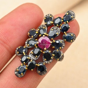 May include: A silver ring with a cluster of blue sapphires and a single red ruby in the center. The ring is set with small diamonds around the sapphires.