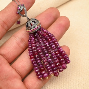 Puede incluir: Un collar de plata y oro con un colgante de borla de rubí. El colgante está adornado con pequeños diamantes y rubíes. La borla está hecha de muchas pequeñas cuentas de rubí.