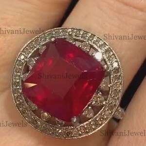 Op de afbeelding: Een opvallende ring met een grote, vierkant geslepen robijn, omringd door een halo van kleinere stenen. De robijn heeft een dieprode kleur en de ring heeft een vintage design. De ring is gezet op een zilveren band.