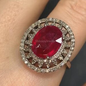 May include: Oval ruby ring with a halo of small diamonds. The ring features a large, faceted red ruby stone set in a silver-coloured metal band. The halo of diamonds surrounds the ruby, enhancing its brilliance. The ring is a statement piece.