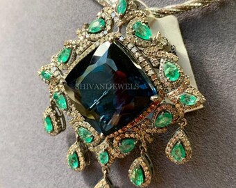 May include: A silver necklace with a large, dark blue gemstone pendant surrounded by smaller green gemstones. The pendant is set in a silver frame with a delicate design. The necklace is on a gray background.