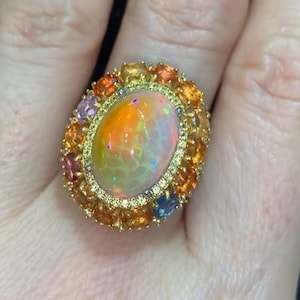 Op de afbeelding: Een gouden ring met een grote ovale opaal omringd door een halo van kleinere edelstenen in verschillende kleuren, waaronder oranje, geel, roze en blauw.