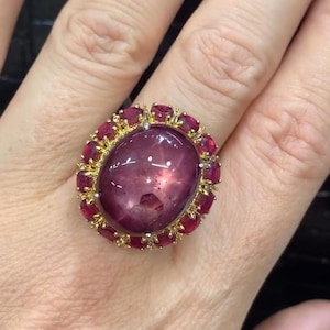 May include: A large, ornate ring featuring a large, oval-shaped, purplish-red gemstone as the centerpiece. The gemstone is surrounded by smaller, oval-cut red gemstones and a gold-colored setting. The ring is a statement piece.