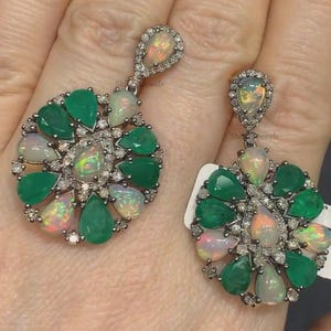 May include: A pair of dangle earrings featuring a round cluster of green and white gemstones. The earrings have a pear-shaped opal gemstone in the centre of the cluster, surrounded by smaller emerald and diamond gemstones.