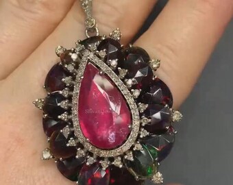 May include: A teardrop-shaped pendant featuring a large red gemstone surrounded by smaller black and green gemstones and diamonds. The pendant is set in silver.