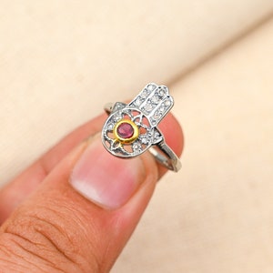 May include: Silver-toned ring featuring a Hamsa hand design. The hand is adorned with small, clear stones and a central gold-colored circle holding a red gemstone. The ring is held against a neutral background.