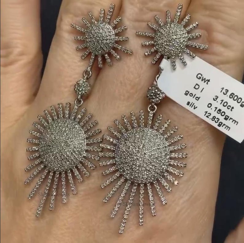 May include: A pair of silver earrings with a sunburst design. Each earring features a large, round, pave-set diamond sun with smaller diamond sunbursts hanging below. The earrings are 13.600g in weight, with 3.10ct of diamonds and 12.83g of silver.