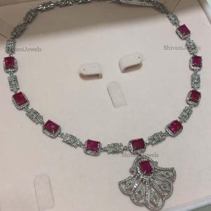 Pode incluir: Um colar de prata com um pingente em forma de leque adornado com diamantes brancos e um grande rubi vermelho no centro. O colar &eacute; adornado com rubis vermelhos menores e diamantes brancos.