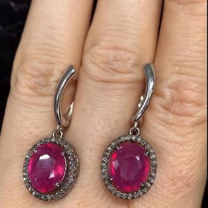 Puede incluir: Un par de pendientes de plata con un gran rubí ovalado rodeado por un halo de diamantes más pequeños.