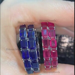 May include: Two gemstone rings, one with blue stones and the other with red stones. Each ring features a series of square-cut stones. The rings are held up by a person's hand. The text "Shivani Jewels" is visible on the image.