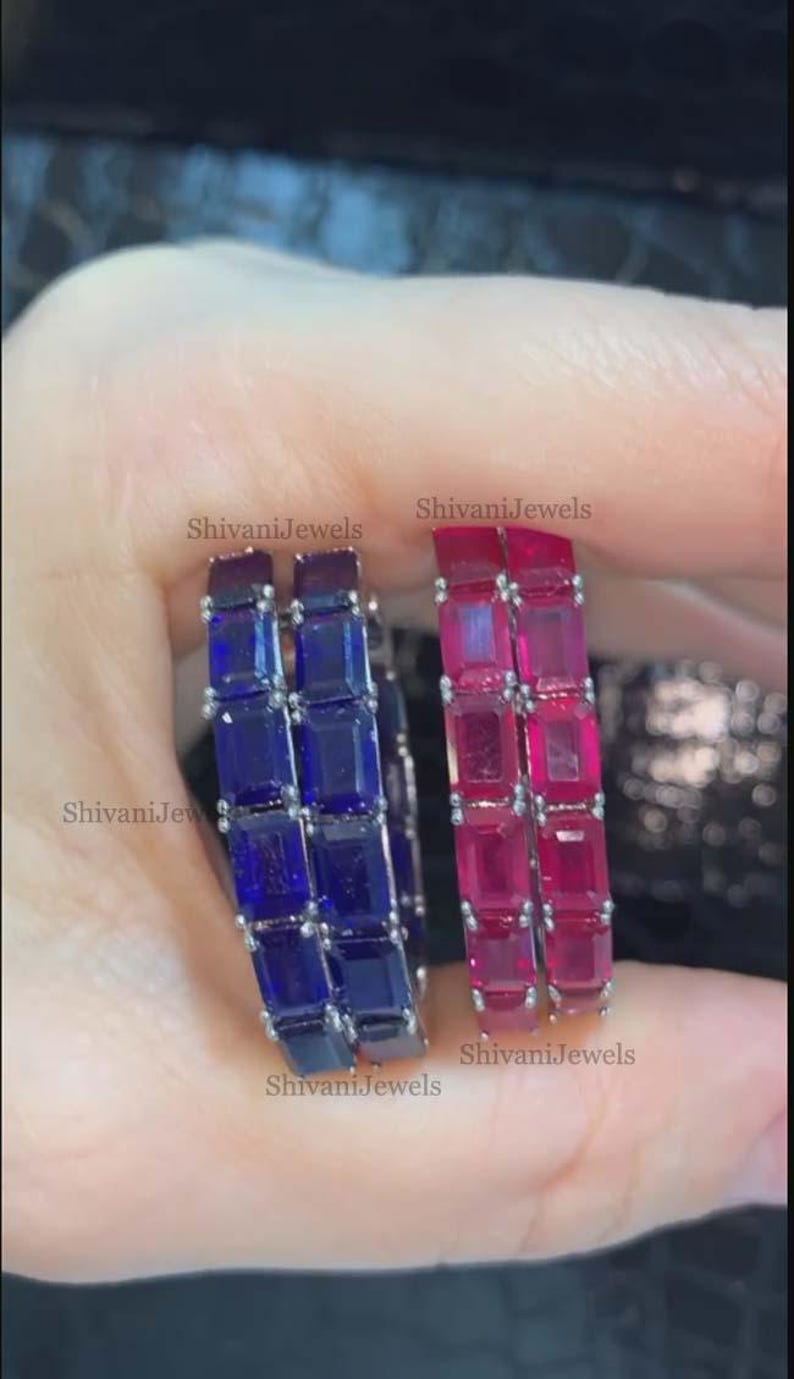 May include: Two gemstone rings, one with blue stones and the other with red stones. Each ring features a series of square-cut stones. The rings are held up by a person's hand. The text "Shivani Jewels" is visible on the image.