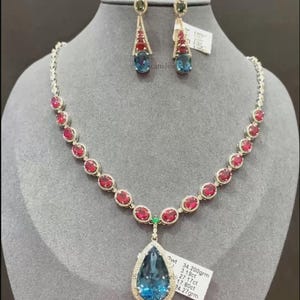 May include: A necklace and earring set featuring a teardrop-shaped blue gemstone pendant and matching blue and red gemstones. The necklace has a series of red gemstones. The earrings have a similar design. The jewelry is displayed on a gray mannequin head.