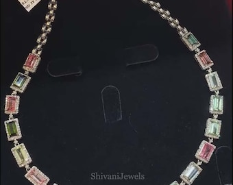 May include: A delicate necklace featuring a series of rectangular gemstones in shades of pink, green, and blue, set in a silver chain. Each gemstone is surrounded by smaller, clear stones. The necklace is displayed on a black surface.