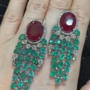 May include: A pair of dangle earrings featuring a large red gemstone surrounded by smaller white gemstones. The earrings are made of silver and have a cascade of green gemstones.