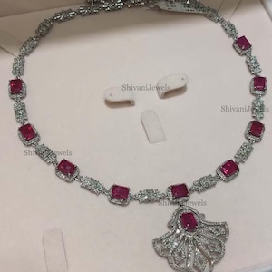 Pode incluir: Um colar de prata com um pingente em forma de leque. O colar &eacute; adornado com pedras preciosas vermelhas e pedras de zirc&ocirc;nia c&uacute;bica transparentes. O pingente tamb&eacute;m &eacute; decorado com pedras de zirc&ocirc;nia c&uacute;bica transparentes. O colar &eacute; apresentado em uma superf&iacute;cie branca. ShivaniJewels est&aacute; impresso na superf&iacute;cie.