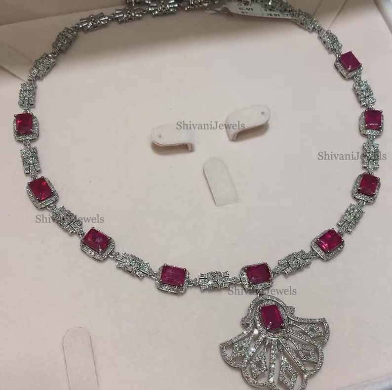 Pode incluir: Um colar de prata com um pingente em forma de leque. O colar &eacute; adornado com pedras preciosas vermelhas e pedras de zirc&ocirc;nia c&uacute;bica transparentes. O pingente tamb&eacute;m &eacute; decorado com pedras de zirc&ocirc;nia c&uacute;bica transparentes. O colar &eacute; apresentado em uma superf&iacute;cie branca. ShivaniJewels est&aacute; impresso na superf&iacute;cie.