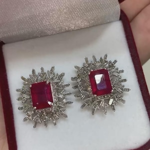 Op de afbeelding: Een paar zilveren oorbellen met een rode edelsteen in het midden van elke oorbel. De oorbellen zijn omgeven door kleinere, heldere edelstenen.