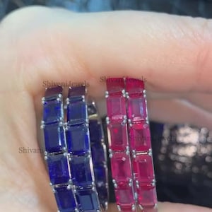 May include: Two silver hoop earrings, one with deep blue rectangular stones and the other with bright pink rectangular stones. The earrings are held up by a hand. The text "Shivani Jewels" is visible on the earrings.