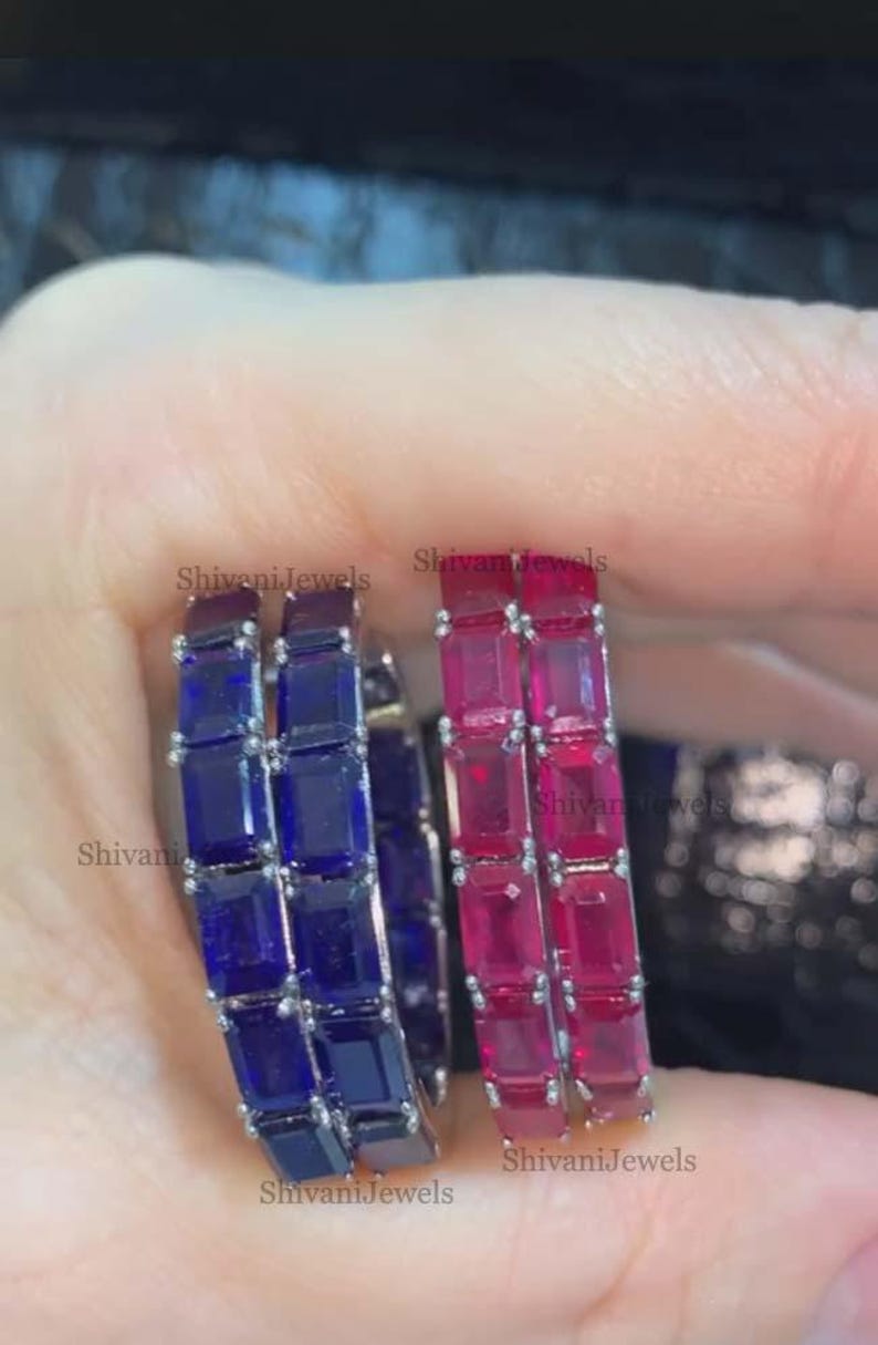 May include: Two silver hoop earrings, one with deep blue rectangular stones and the other with bright pink rectangular stones. The earrings are held up by a hand. The text "Shivani Jewels" is visible on the earrings.