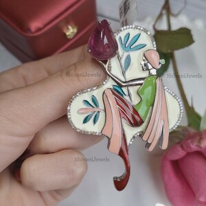 May include: A colorful brooch featuring a stylized figure holding a large, teardrop-shaped, deep pink gemstone. The brooch has a white background with teal and red accents, and is outlined with small, clear stones. The text "ShivaniJewels" is visible.