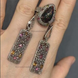 May include: A silver ring and matching earrings set. The ring features a large, pear-shaped black gemstone surrounded by smaller, colorful gems. The earrings are rectangular with a similar design, showcasing a mix of pink, yellow, and clear stones. The jewelry is set against a neutral background.