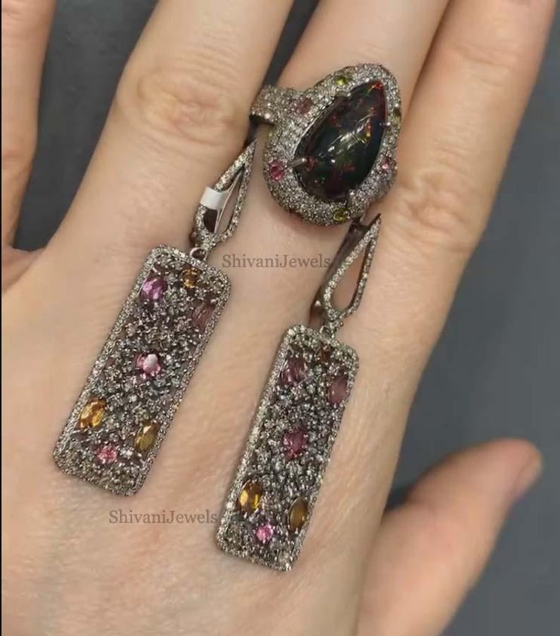 May include: A silver ring and matching earrings set. The ring features a large, pear-shaped black gemstone surrounded by smaller, colorful gems. The earrings are rectangular with a similar design, showcasing a mix of pink, yellow, and clear stones. The jewelry is set against a neutral background.