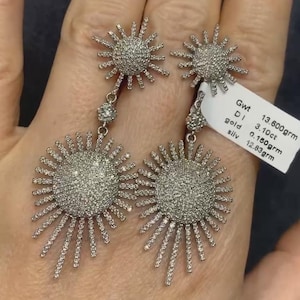 May include: A pair of silver dangle earrings with a sunburst design. Each earring features a round center with a pave setting of small diamonds. The earrings are 13.60 grams, 3.10 carats of diamonds, 0.180 grams of gold, and 12.83 grams of silver.