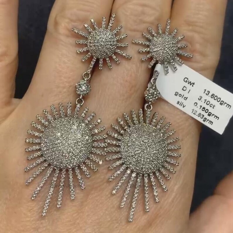 May include: A pair of silver dangle earrings with a sunburst design. Each earring features a round center with a pave setting of small diamonds. The earrings are 13.60 grams, 3.10 carats of diamonds, 0.180 grams of gold, and 12.83 grams of silver.