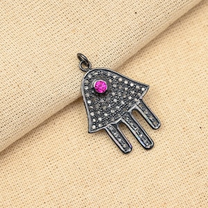 May include: A silver Hamsa hand charm pendant with a pink gemstone in the center and black diamonds surrounding it.