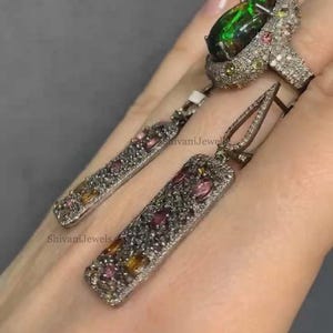 May include: A silver ring and matching earrings set. The ring features a large, oval, dark green gemstone. The earrings and ring are encrusted with small, colorful gemstones. The text "Shivani Jewels" is visible.