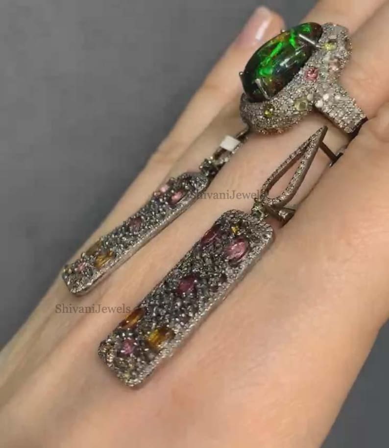 May include: A silver ring and matching earrings set. The ring features a large, oval, dark green gemstone. The earrings and ring are encrusted with small, colorful gemstones. The text "Shivani Jewels" is visible.