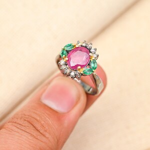 May include: A silver ring featuring a large oval ruby gemstone, surrounded by smaller diamonds and green emeralds.