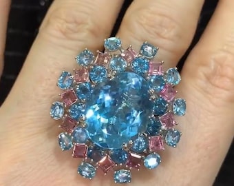 May include: A large, oval-shaped blue topaz gemstone ring surrounded by smaller blue and pink gemstones. The ring is set in silver and has a floral design. The text "SHIVANI JEWELS" is visible below the ring.