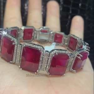 Può includere: Un braccialetto d'argento con una fila di pietre preziose rettangolari rosse incastonate in argento. Le pietre preziose sono circondate da pietre preziose trasparenti più piccole. Il braccialetto è al polso di una persona.