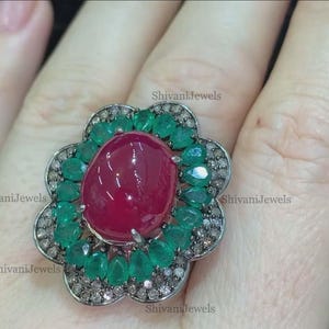 May include: A silver ring featuring a large red gemstone encircled by green gemstones and smaller white gemstones. The ring is shaped like a flower.