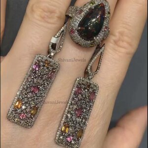 May include: A silver ring and matching earrings set. The ring features a large, dark gemstone surrounded by smaller gems. The earrings are rectangular with colorful gems. The jewelry is set against a neutral background.