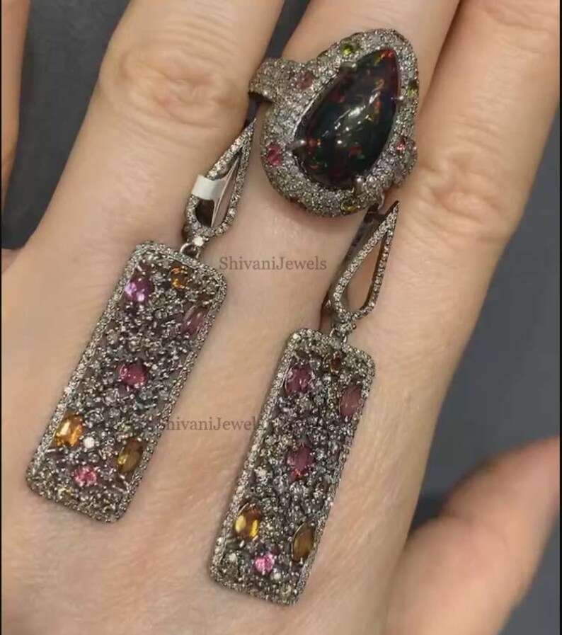 May include: A silver ring and matching earrings set. The ring features a large, dark gemstone surrounded by smaller gems. The earrings are rectangular with colorful gems. The jewelry is set against a neutral background.